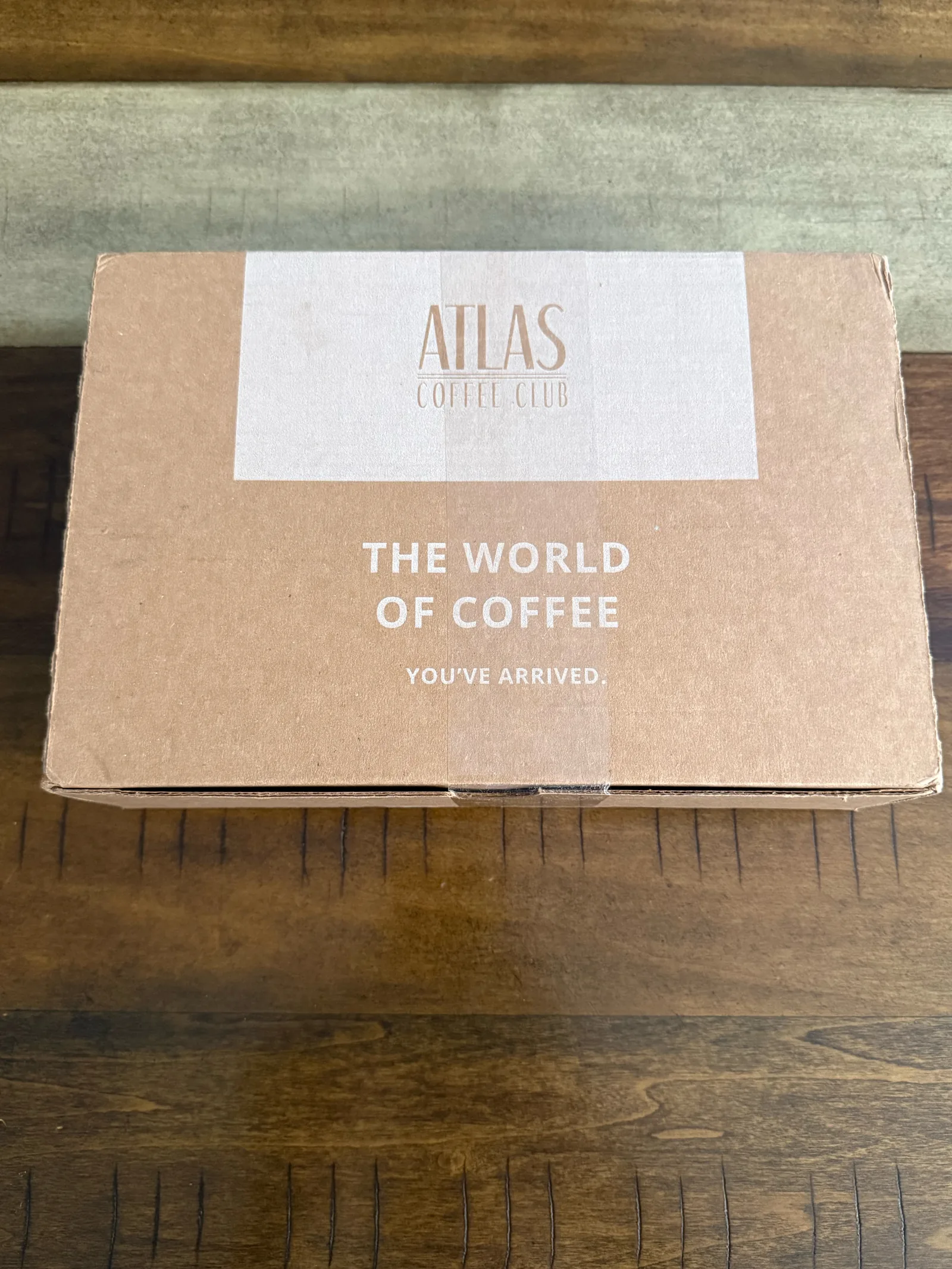 Closed Atlas Coffee Club shipping box reading "The world of coffee. You've arrived." on a wooden table