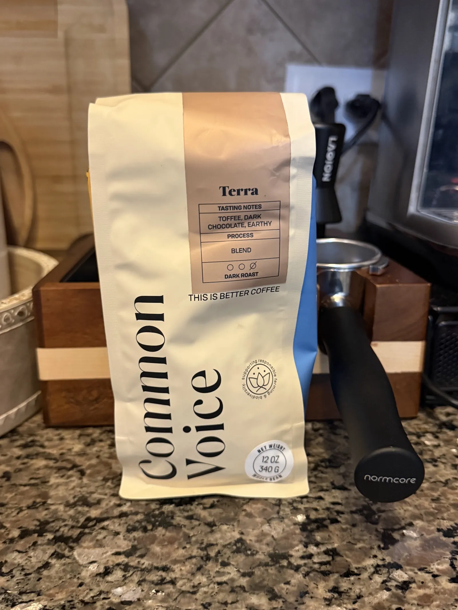 Common Voice "Terra" coffee bag standing on a granite counter next to a wooden knock box and a Normcore portafilter