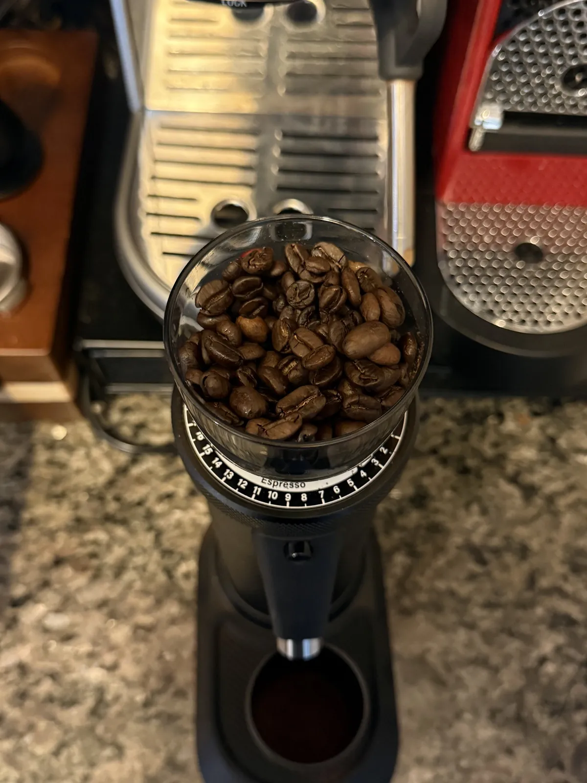 Top-down view of whole coffee beans in a grinder hopper, the dial marked "Espresso", with an espresso machine behind