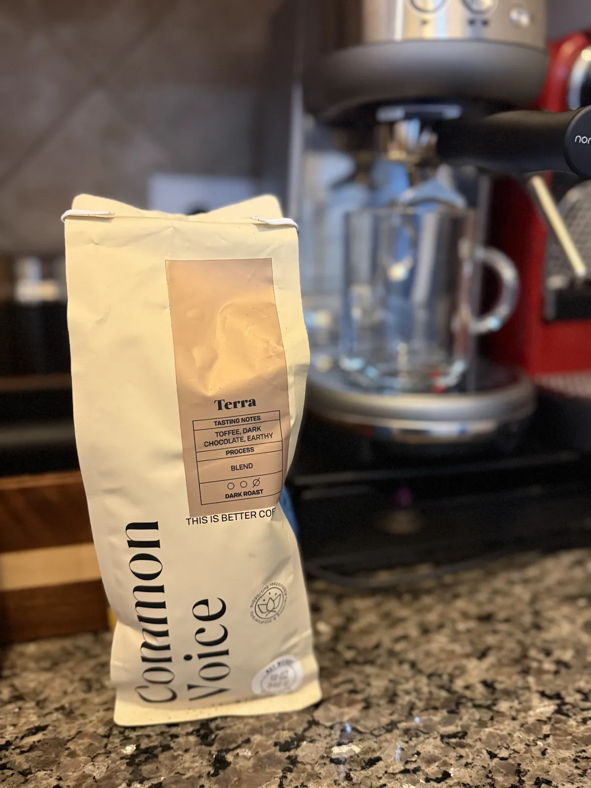 Common Voice "Terra" bag on a granite counter in front of an espresso machine with an empty glass mug under the spout