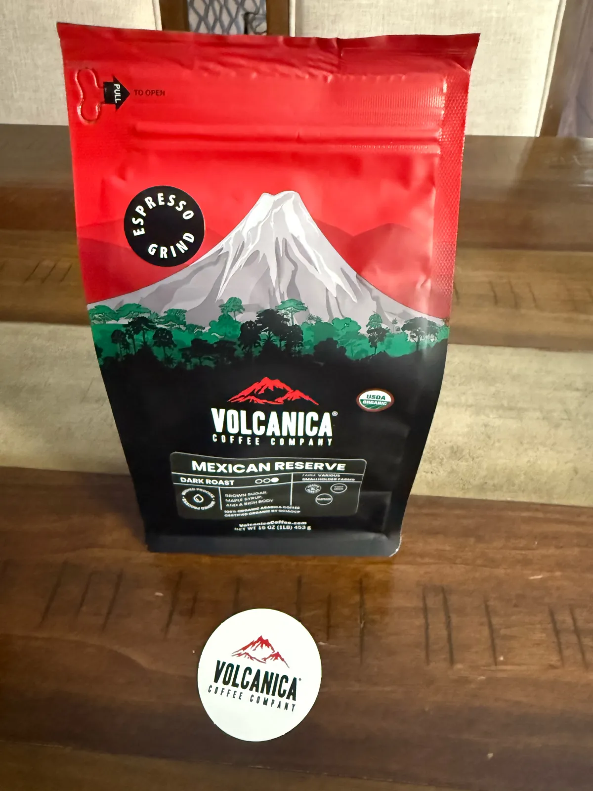 Front of the red Volcanica Mexican Reserve bag with a volcano illustration and USDA Organic seal, next to a round Volcanica sticker